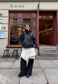 Veste en cuir noir, robe blanche asymétrique oversize avec bordure en dentelle, pantalon large noir, chaussures noires épaisses, tenant une tasse noire.