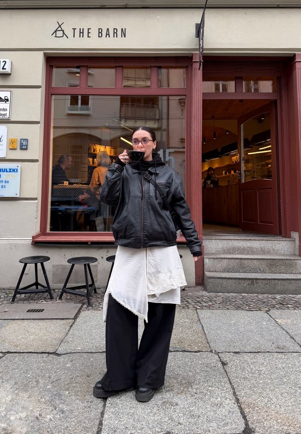 Blouson en cuir noir, robe blanche asymétrique surdimensionnée avec bordure en dentelle, pantalon noir large et chaussures noires épaisses, tenant une tasse noire.