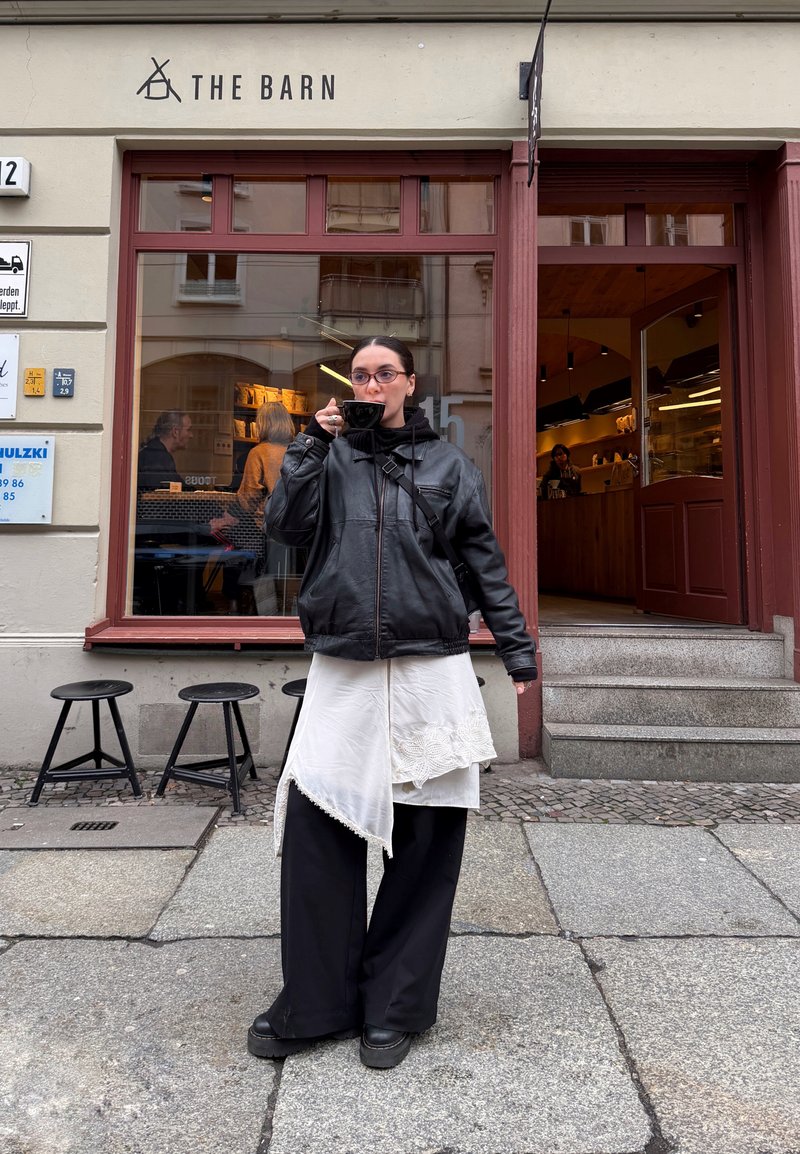Veste en cuir noir, robe blanche asymétrique oversize avec bordure en dentelle, pantalon large noir, chaussures noires épaisses, tenant une tasse noire.