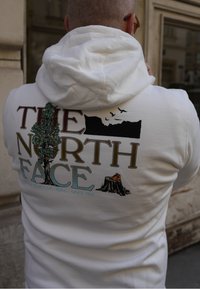 Weisse Kapuzenjacke mit "The North Face"-Logo. Das grafische Design zeigt einen Baum, Berge und orangefarbene Schuhe auf einem Baumstumpf. Mit Kapuze.