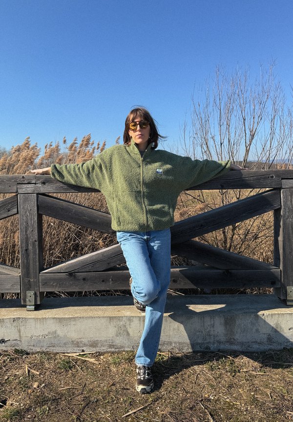 Personne portant une veste polaire verte et des lunettes de soleil, appuyée contre une clôture en bois avec une jambe croisée, sous un ciel bleu clair.