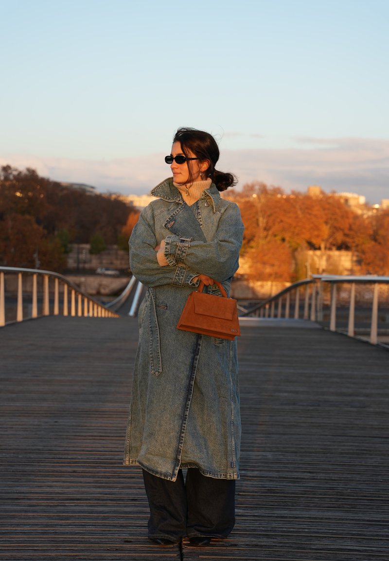 Trench-coat en denim avec un col montant, une coupe oversized et des fentes latérales. Sac à main marron slim et lunettes de soleil noires complètent le look.