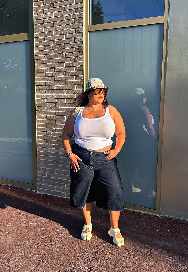 Femme portant un débardeur blanc, un pantalon large noir, un bonnet beige en tricot et des lunettes de soleil, debout contre un mur en briques et en verre sous la lumière du soleil.