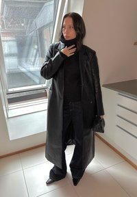 Černý kožený dvouřadý kabát přes černý rolák, spárovaný s rozšířenými tmavými džíny a černými botami s špičatou špičkou.