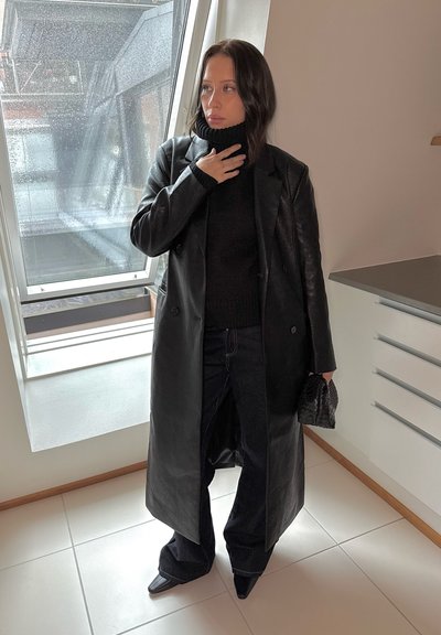 Abrigo de cuero negro de doble botonadura sobre un sweater negro de cuello alto, combinado con jeans de mezclilla oscuros acampanados y botas negras de puntera afilada.