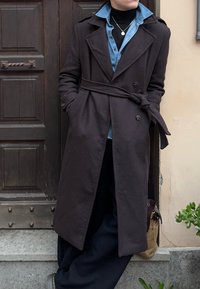 Personne appuyée contre une porte en bois, portant un manteau sombre ceinturé par-dessus une chemise en denim et un col roulé noir, avec un petit pendentif visible.