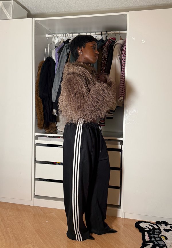 Jeune personne portant une veste en fausse fourrure et un pantalon noir à rayures blanches, debout devant une armoire ouverte remplie de vêtements.