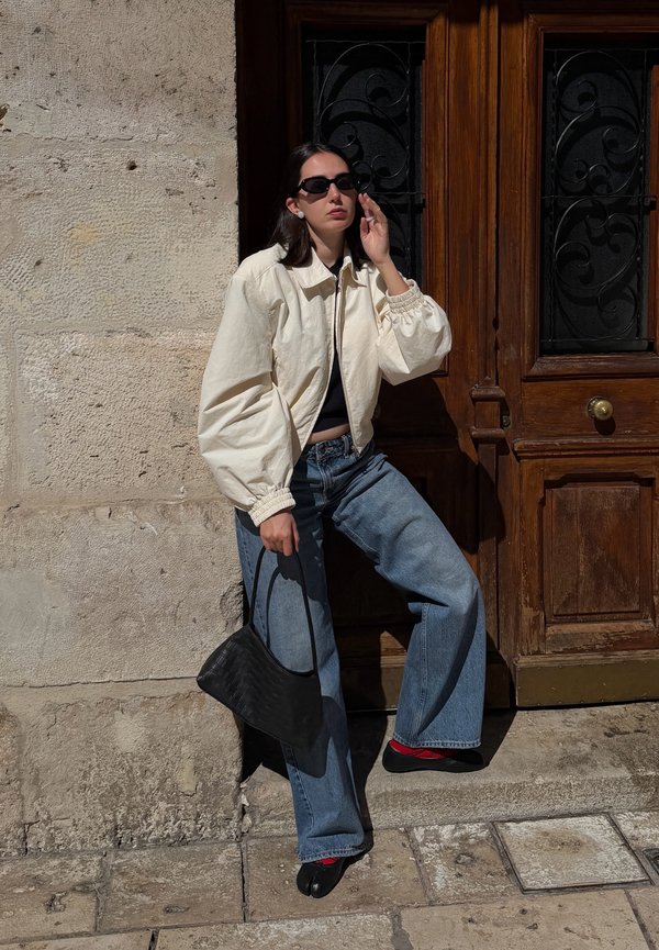 Mujer con gafas de sol que lleva una chaqueta crema y vaqueros anchos, está de pie junto a un muro de piedra, sosteniendo un bolso negro cerca de una puerta de madera.