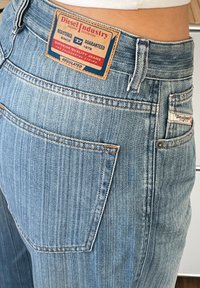 Calças de ganga azul desbotadas com um bolso traseiro cosido, apresentando uma etiqueta de marca em pele com accents em vermelho e azul na cintura.