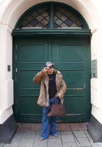 Kabát z umělé kožešiny v odstínech hnědé, černé tričko, rozšířené džíny z denimu, hnědá semišová kabelka, sluneční brýle, stojí u zelených dveří.