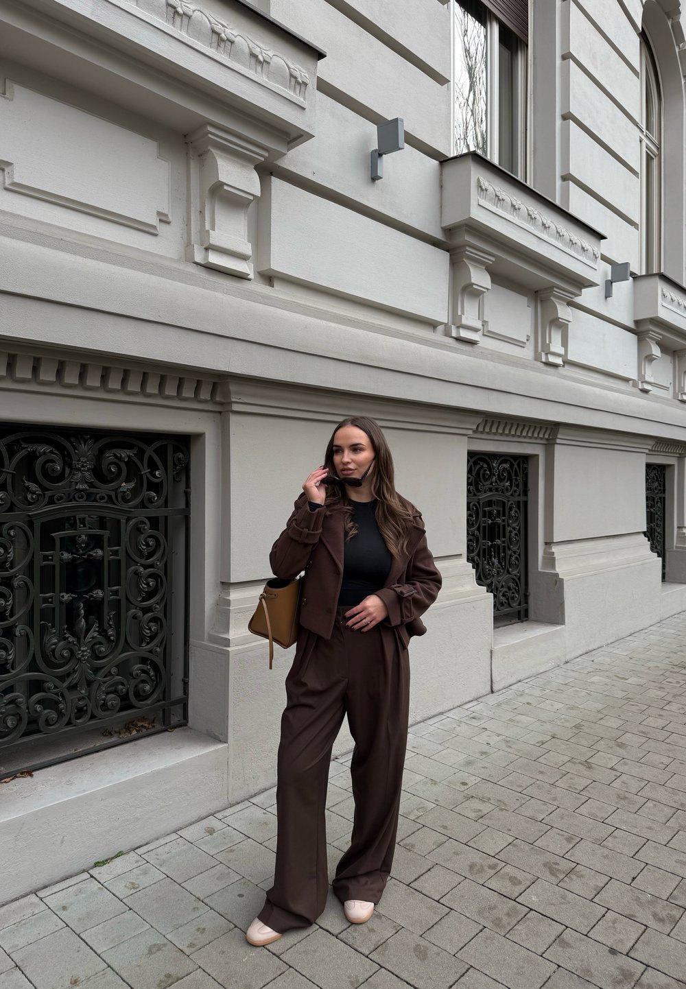 Καφέ κοστούμι κομμένο με ανοιχτές παντελόνες και εφαρμοστό τοπάκι. Λείο υφασμά, απαλός φωτισμός. Καφέ τσάντα, ανοιχτόχρωπα παπούτσια και περίτεχνα σιδερένια στοιχεία στο φόντο.