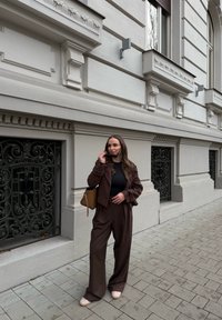 Καφέ κοστούμι κομμένο με ανοιχτές παντελόνες και εφαρμοστό τοπάκι. Λείο υφασμά, απαλός φωτισμός. Καφέ τσάντα, ανοιχτόχρωπα παπούτσια και περίτεχνα σιδερένια στοιχεία στο φόντο.