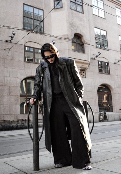 Persona con abrigo de cuero negro y pantalones anchos apoyada en un soporte metálico para bicicletas en una calle de la ciudad con un edificio de piedra y ventanas detrás.