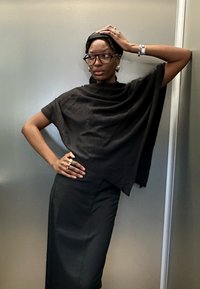 Čierny, textúrovaný, skrátený pončo vrch s dlhou, priliehavou čiernou sukňou. Modelka nosí okuliare, prstene a výrazné náušnice.