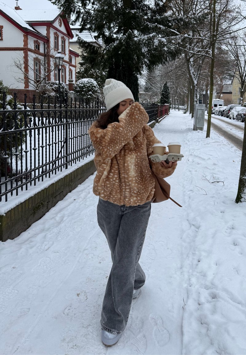 Persoană cu căciulă albă tricotată și jachetă de blană artificială maro cu pete, ținând două căni de cafea și mergând pe o trotuar acoperit de zăpadă, mărginit de copaci și mașini.