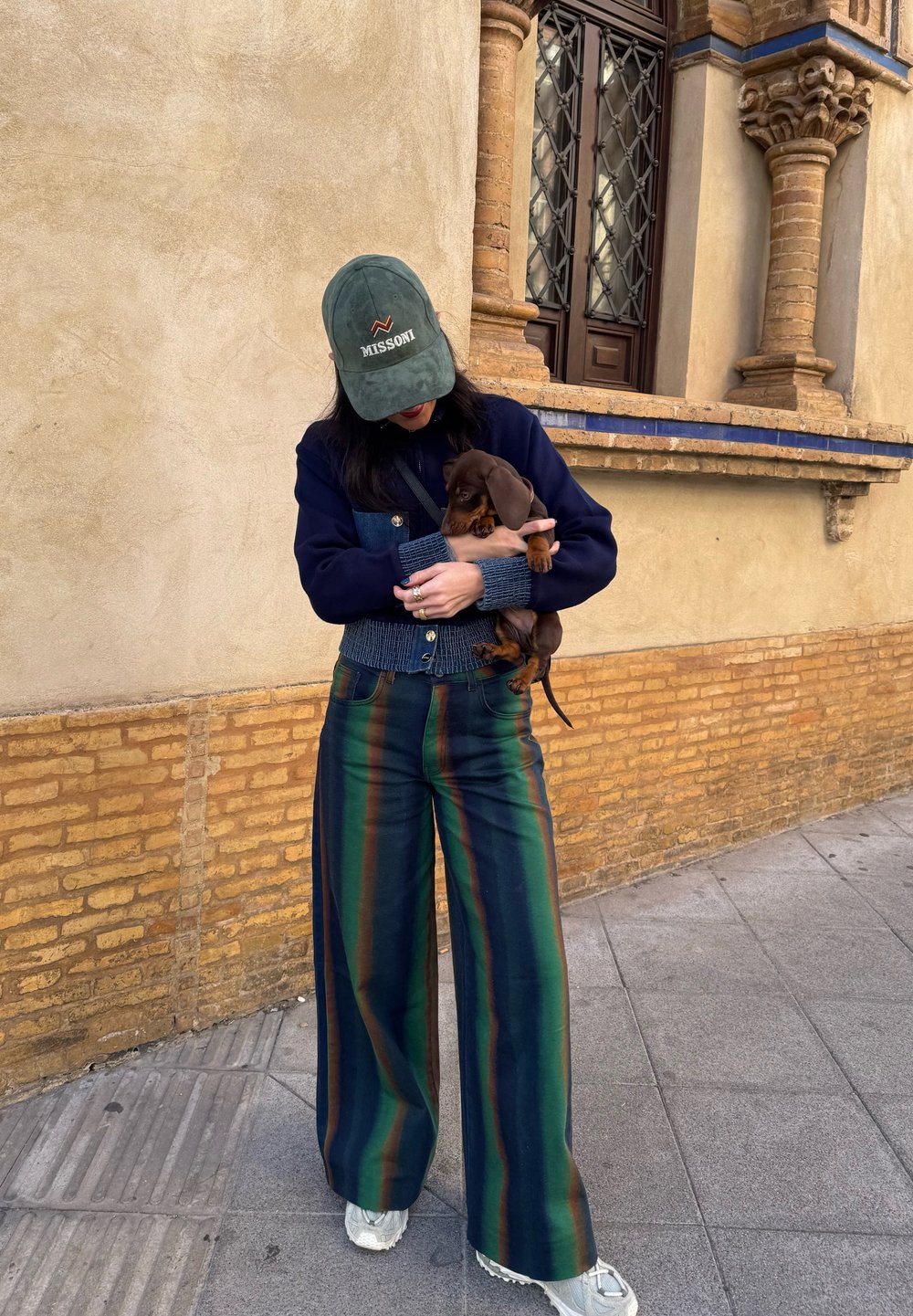 Τζιν μπλούζα με μπλε λεπτομέρειες, πράσινο καπέλο, πολυχρωμοί ριγωτοί παντελόνες σε σκούρο πράσινο, ναυτικό και καφέ. Κρατάει ένα καφέ κουτάβι.