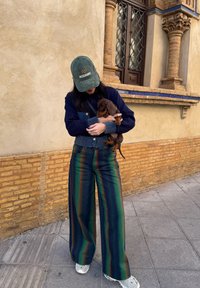 Τζιν μπλούζα με μπλε λεπτομέρειες, πράσινο καπέλο, πολυχρωμοί ριγωτοί παντελόνες σε σκούρο πράσινο, ναυτικό και καφέ. Κρατάει ένα καφέ κουτάβι.