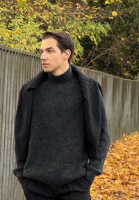 Tumšs adīts džemperis ar augstu apkakli, saskaņots ar melnu virsjaku. Svītrainas melnas bikses, stāvot lapu apvemtā rudenīgā dārza vidē.