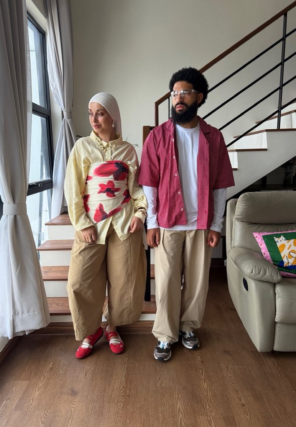 Due persone stanno dentro vicino alle scale e alla finestra; una indossa un foulard e un top floreale, l'altra porta occhiali e una camicia rossa a quadri.