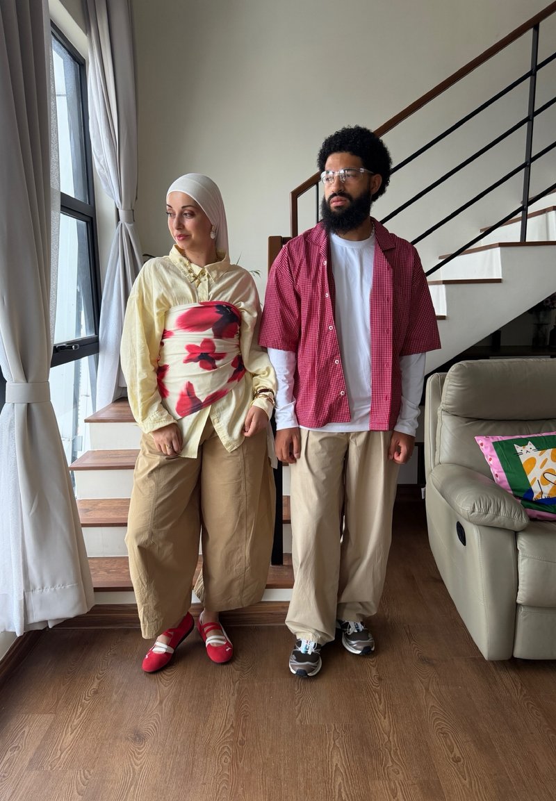 Deux personnes se tiennent à l'intérieur près des escaliers et d'une fenêtre ; l'une porte un foulard et un haut fleuri, l'autre porte des lunettes et une chemise à carreaux rouge.