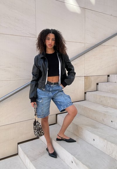 Chaqueta bomber negra corta, top negro corto, pantalones cortos de denim con cargo y zapatos negros tipo slingback. Lleva un bolso de leopardo. El suelo es de piedra clara.