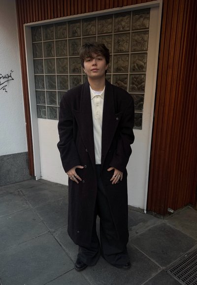 Abrigo largo negro con mangas anchas, camisa polo blanca, pantalones negros de gran tamaño, zapatos negros, anillos plateados, de pie frente a un fondo texturizado.