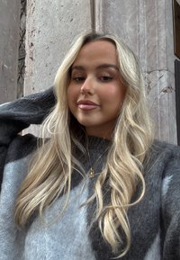 Jeune femme aux longs cheveux blonds portant un pull gris, posant avec la main derrière la tête devant un mur de pierre.