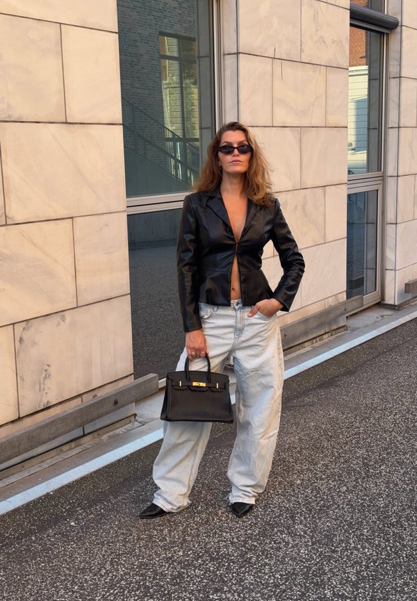 Femme portant une veste en cuir noire, un jean ample clair et des lunettes de soleil noires, debout sur un trottoir tenant un sac à main noir.