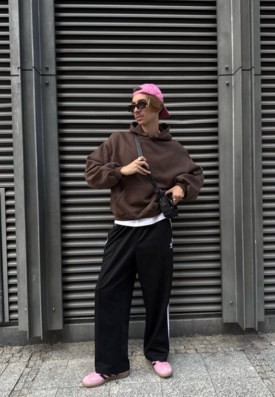 Joven con una sudadera marrón, pantalones Adidas negros, zapatillas rosas, gorra rosa al revés y gafas de sol, de pie contra una pared de ventilación metálica.