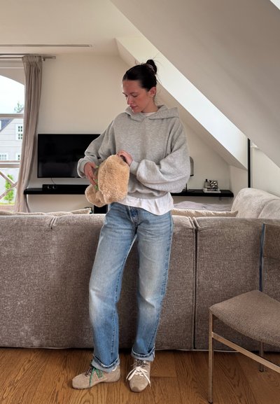 Sudadera oversized gris claro, jeans azules y zapatillas beige. Sosteniendo un bolso de peluche marrón en una habitación neutra con un sofá y una televisión.