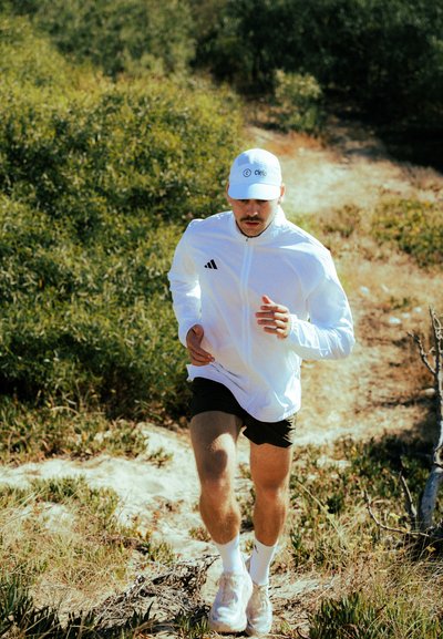 Chaqueta ligera blanca con cremallera, pantalones cortos negros y una gorra blanca. El corredor está en un sendero natural con follaje verde de fondo.