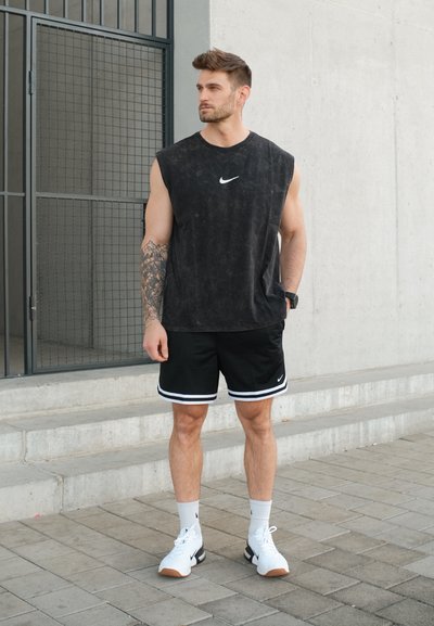 Hombre con camiseta negra sin mangas de Nike, pantalones cortos negros, calcetines blancos y zapatillas blancas de pie sobre un pavimento de baldosas cerca de unos escalones de hormigón.