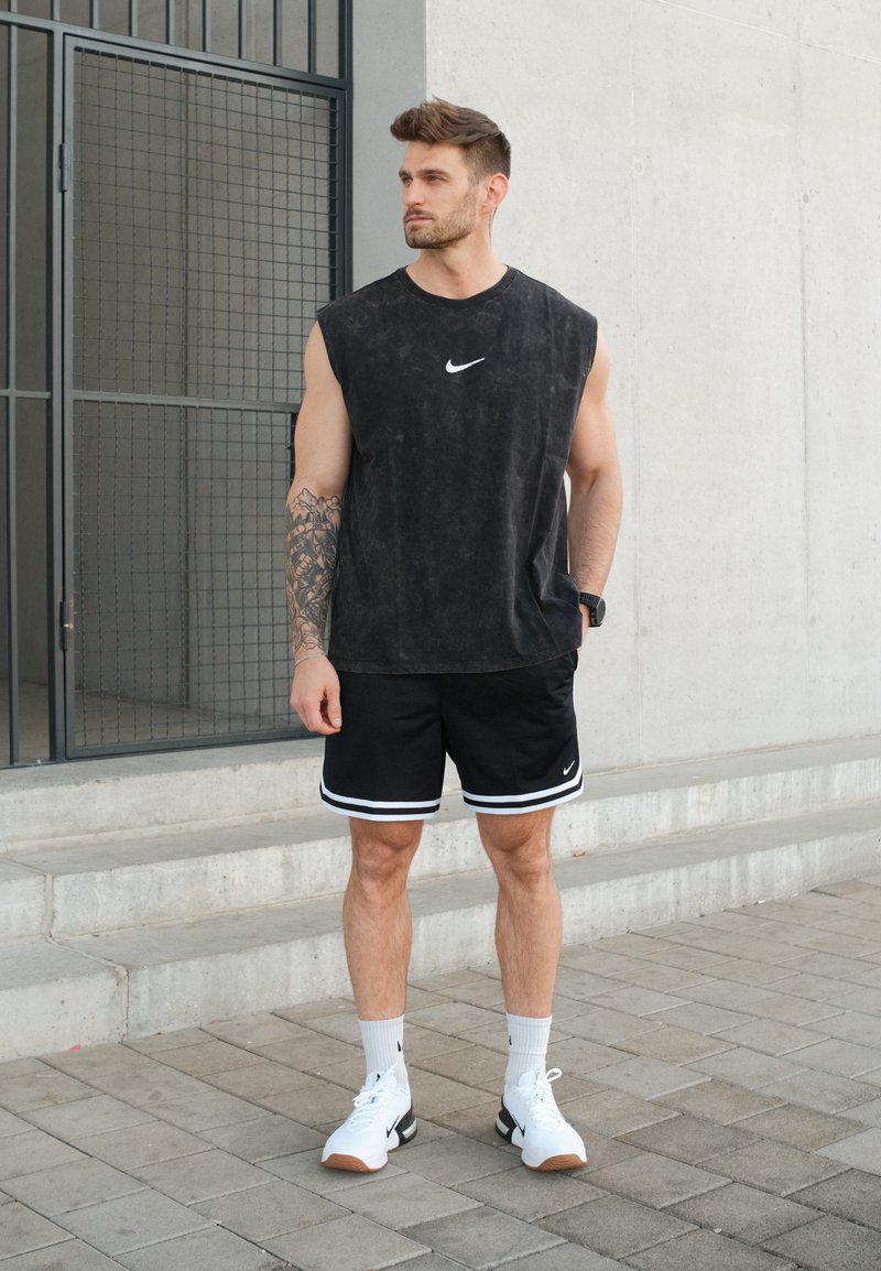 Homme portant un débardeur Nike noir, un short noir, des chaussettes blanches et des baskets blanches, debout sur un trottoir pavé près d'escaliers en béton.