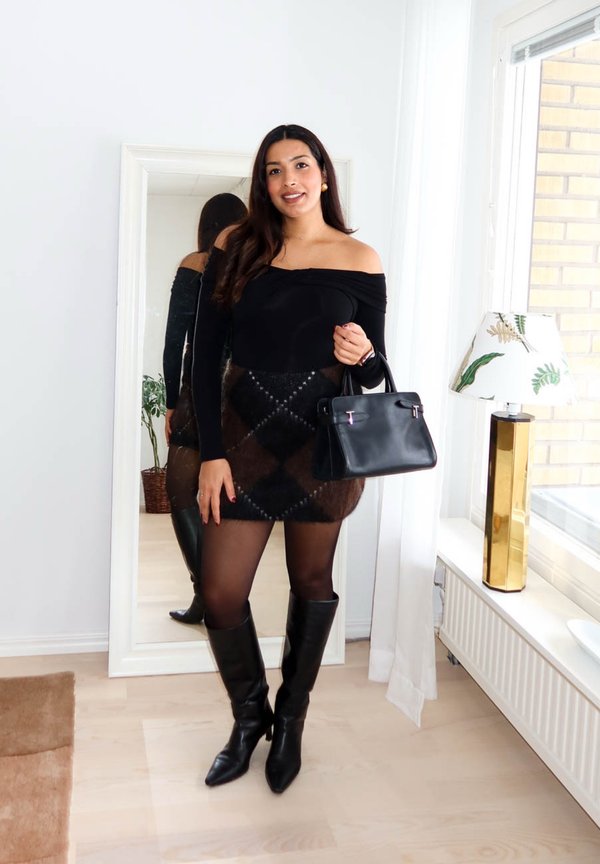 Sieviete valkā melnu vaļēju plecu topu, rūtainu brūnu svārku, melnas ceļgalu zābakus, tur rokā melnu rokassomu, stāvot priekšā pilna auguma spogulim.