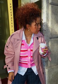 Różowy płaszcz trencz na białej koszuli i różowym swetrze w paski, zestawiony z spodniami w paski. Trzyma filiżankę kawy, prezentując kolorowy manicure.