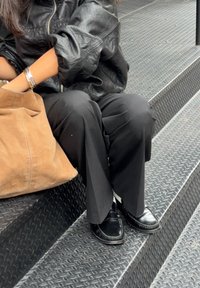Una chaqueta de cuero negra, pantalones de pierna ancha negros, mocasines negros y un bolso de ante marrón reposan sobre unos escalones de metal con superficie texturada.