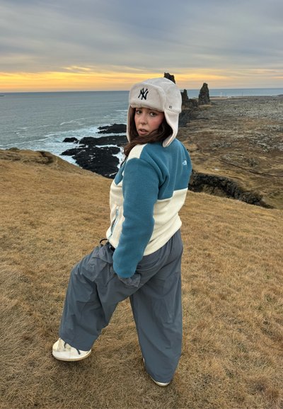 Persona con gorro y chaqueta de invierno mirando hacia atrás mientras está de pie en un acantilado cubierto de hierba cerca del océano al atardecer, con formaciones rocosas en el fondo.