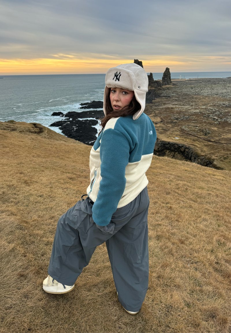 Person mit Wintermütze und Jacke blickt zurück, während sie auf einer grasbewachsenen Klippe nahe dem Ozean bei Sonnenuntergang mit felsigen Formationen im Hintergrund steht.