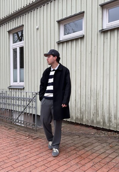 Joven con abrigo negro, suéter a rayas, pantalones grises y gorra caminando por una acera de ladrillo junto a un edificio de madera verde pálido.