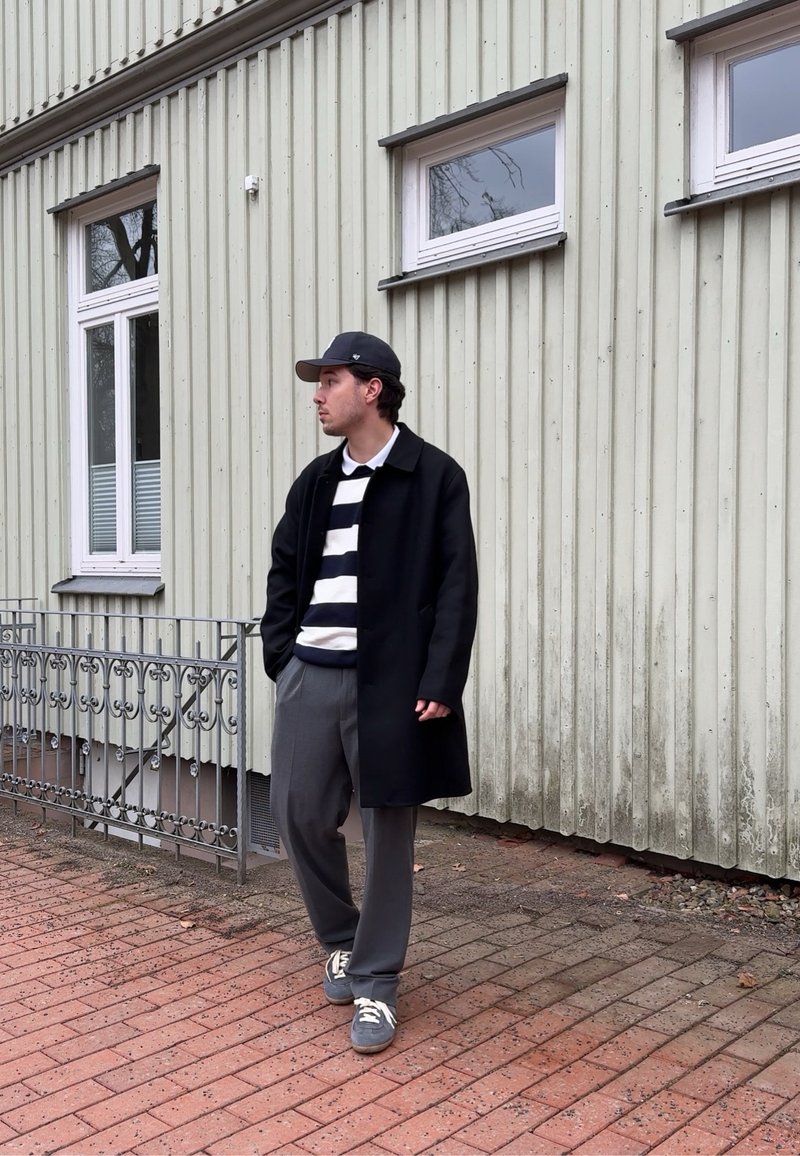 Jeune homme portant un manteau noir, un pull rayé, un pantalon gris et une casquette, marchant sur un trottoir en briques à côté d'un bâtiment en bois vert pâle.
