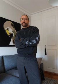 Prillidega ja vuntsidega mees, kellel on seljas tume dressipluus ja triibulised püksid, seisab kaasaegses elutoas käed ristatud.