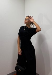 Femme en tenue noire posant contre un mur d'angle blanc, tenant un sac à main noir, la main levée près de son front.