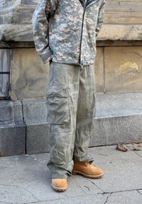 Persona, kas valkā maskēšanās jaku, olīvu krāsas darba bikses un bēšas darba zābakus, stāv uz plaisātas asfalta malas pie akmens sienas.