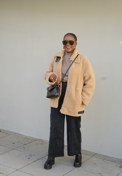 Chaqueta de forro polar beige con cuello, llevando un suéter marrón claro y pantalones de pierna ancha negros, complementada con un pequeño bolso negro y gafas de sol.