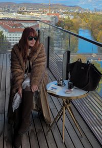 Barna műszőrme kabát, krémszínű szoknya csipkés szegéllyel, térdig érő barna csizma. Kerek asztal fémes felülettel, kávésbögrével és egy könyvvel.