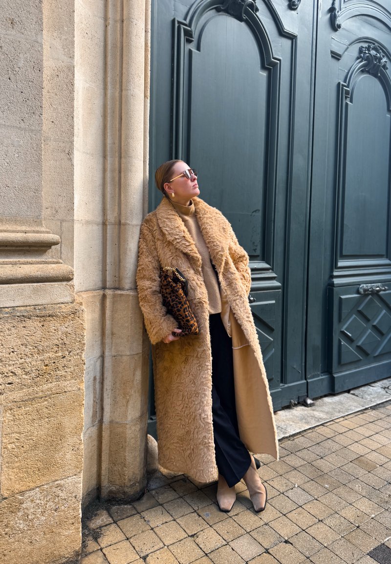Femme en long manteau en fourrure marron, pull beige, pantalon noir et bottes beiges appuyée contre un mur en pierre, tenant une pochette à imprimé léopard près de grandes portes bleues.