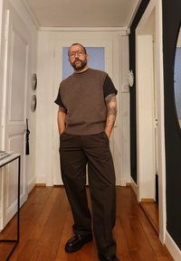 Brązowy dzianinowy top bez rękawów na czarnej koszuli, w zestawie z ciemnobrązowymi spodniami o szerokich nogawkach i czarnymi butami. Drewniana podłoga i białe ściany.