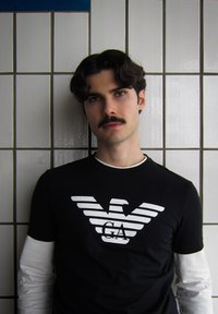 Čierne tričko s bielym pruhovaným logo orla a textom "GA". Dlhé biele rukávy sú viditeľné. Hladká tkanina, okrúhly výstrih, uvoľnený strih.