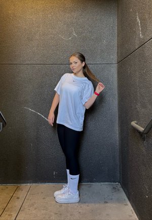 Camiseta blanca de manga corta con un logo, leggings negros, zapatillas blancas y calcetines blancos. De pie contra una pared gris texturizada.