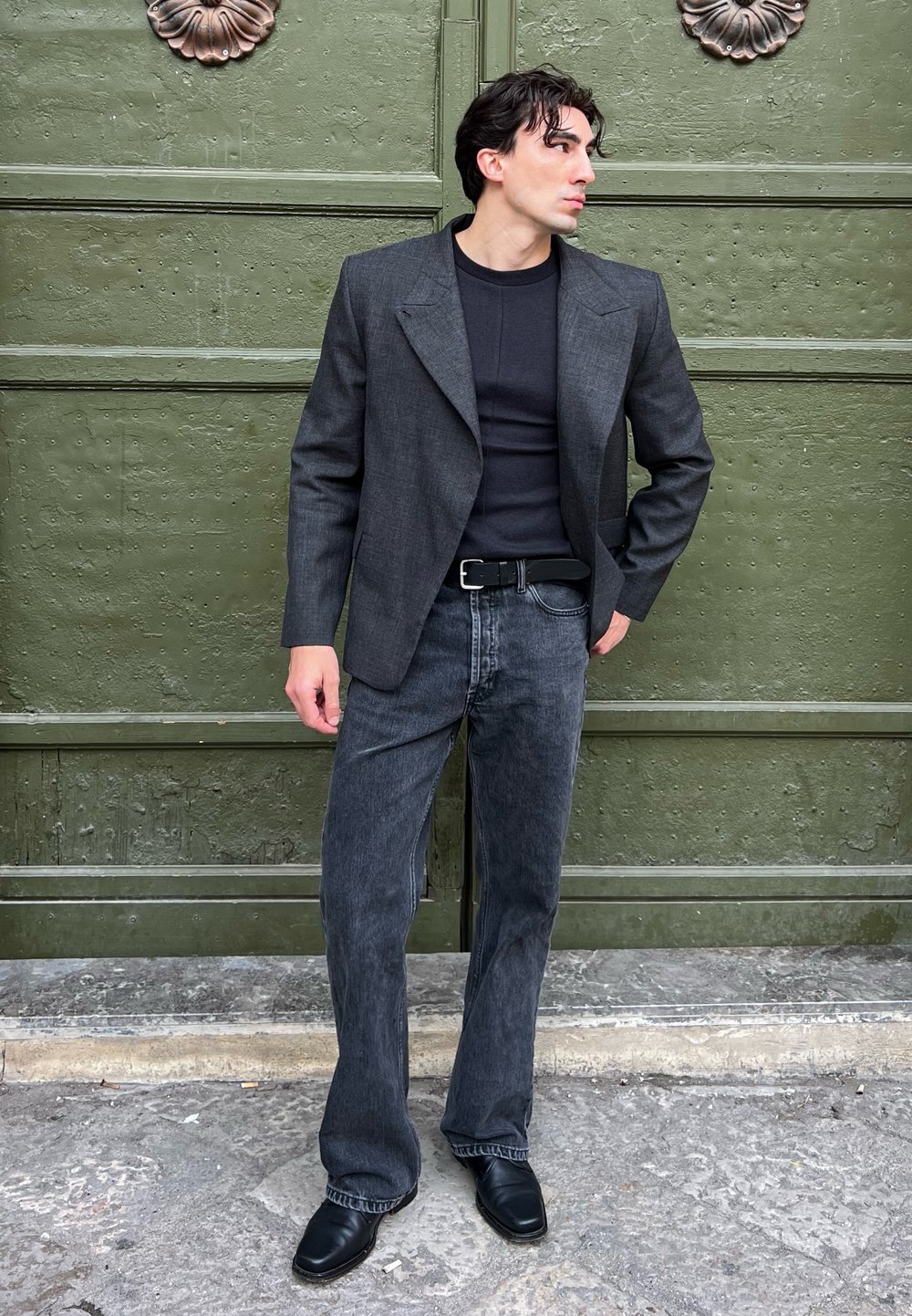 Blazer gris sobre una camiseta negra, combinado con jeans de mezclilla oscura acampanados y zapatos negros. Tela texturizada y diseño estructurado, posando frente a un fondo verde.